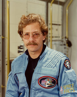 White man with blond short curly hair and a mustache, wearing glasses. He is wearing a blue flight suit with a few patches on it.